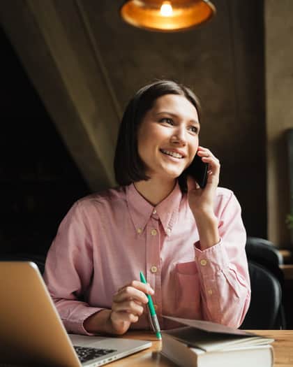 Mulher sorrindo ao telefone, sentada à mesa com um notebook